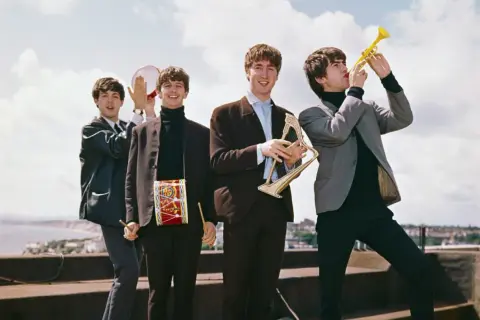 The Beatles w swoich eleganckich garniturach, grając na instrumentach, tworzą radosną atmosferę, jakby zapowiadali nowe utwory.