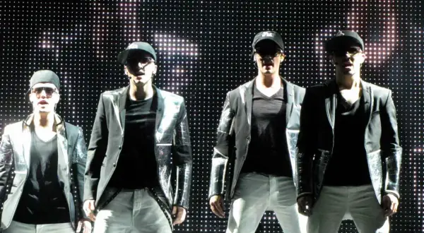 Członkowie Big Time Rush na scenie, w błyszczących marynarkach i czapkach z daszkiem, przed ekranem pełnym świateł.