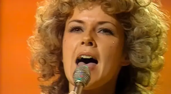 Kobieta z burzą blond loków śpiewa do mikrofonu. Jej makijaż i stylizacja przywodzą na myśl piosenki Abby.
