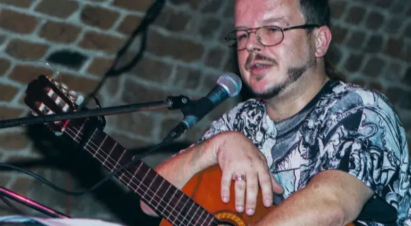 Jacek Kaczmarski z gitarą i mikrofonem, w okularach i wzorzystej koszulce. Prywatne chwile artysty.