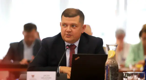 Jacek Wójcicki, siedząc przy stole, przemawia. Jego wzrost jest przeciętny, a na stole leży laptop i woda z cytryną.