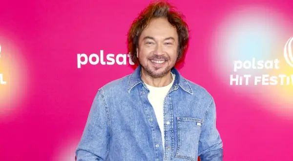 Mietek Szczęśniak, uśmiechnięty, w dżinsowej koszuli, na tle różowego banera z logo Polsat Hit Festiwal.