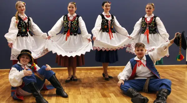 Dzieci w strojach ludowych, dziewczęta trzymają białe spódnice, chłopcy w niebieskich spodniach i kamizelkach, gotowi do tańca kołomyjka.
