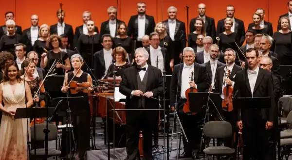 Orkiestra i chór przygotowują się do wykonania **Moniuszko opery**. Dyrygent stoi na podwyższeniu, gotowy do rozpoczęcia.