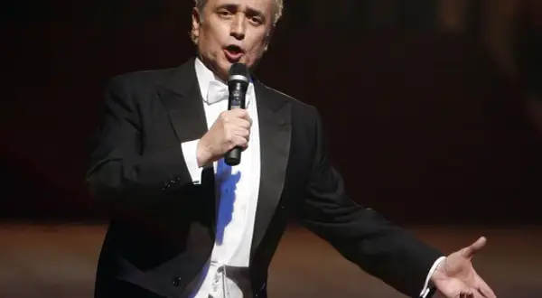 José Carreras, hiszpański tenor, w eleganckim smokingu śpiewa do mikrofonu, gestykulując.