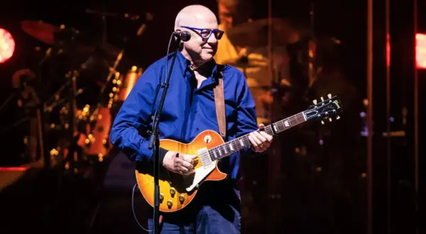 Mark Knopfler, w okularach i niebieskiej koszuli, gra na gitarze Gibson. Jego muzyka, jak na jego albumy, brzmi w tej chwili na żywo.