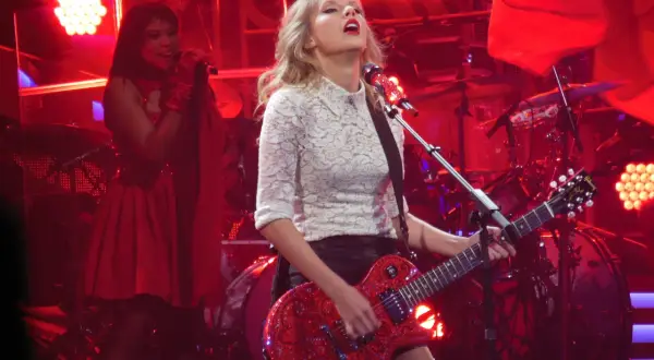 Taylor Swift gra na czerwonej gitarze podczas koncertu. W tle widać perkusję i inną wokalistkę. To może być fragment trasy promującej jej taylor swift albumy.