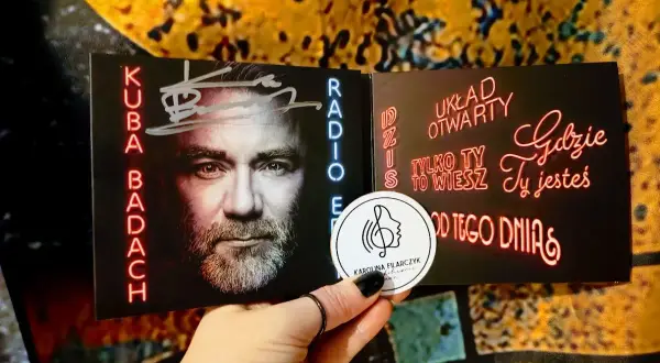 Kuba Badach z autografem na okładce płyty. Neonowe napisy "Dziś układ otwarty", "Tylko ty to wiesz", "Gdzie ty jesteś", "Od tego dnia".