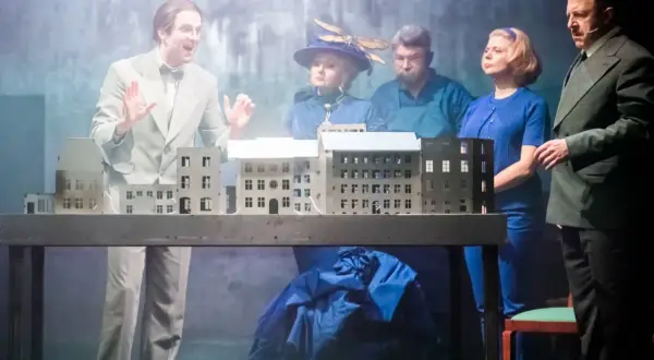 Aktorzy na scenie, prezentujący polskie musicale, gestykulują przy makiecie miasta.