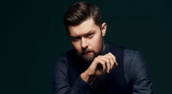 Mężczyzna z brodą i starannie ułożonymi włosami, w ciemnej koszuli i kamizelce, spogląda intensywnie. Jego dłoń opiera się o podbródek, jakby zastanawiał się nad kolejnymi mrozu utworami.
