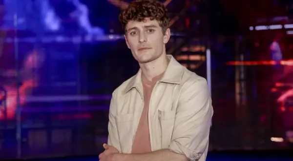 Młody mężczyzna z kręconymi włosami, w beżowej koszuli, stoi na tle sceny. Jakub Józef Orliński wzrostem imponuje, ale jego talent jest jeszcze większy.