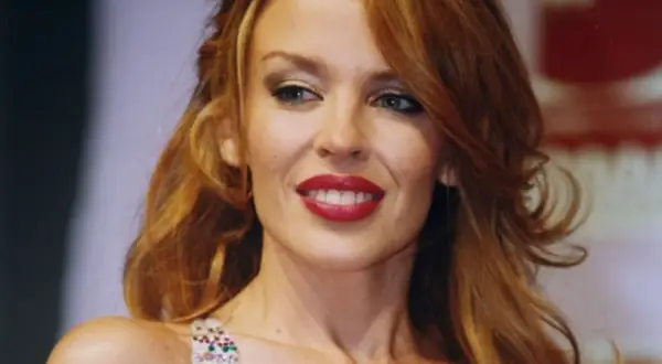 Uśmiechnięta Kylie Minogue, której wiek nie przeszkadza w olśniewaniu. Jej charakterystyczne rude włosy i czerwone usta przyciągają wzrok.