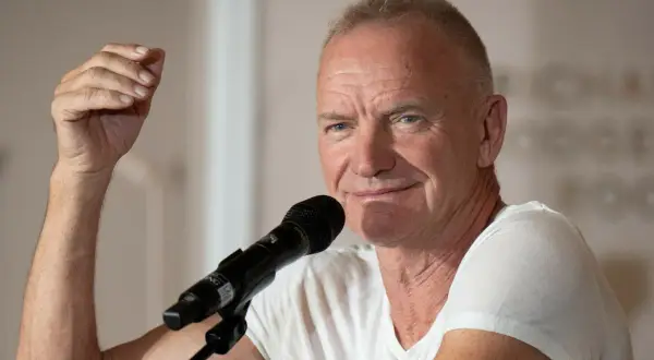 Sting, wokalista zespołu The Police, przemawia do mikrofonu, unosząc rękę w geście.
