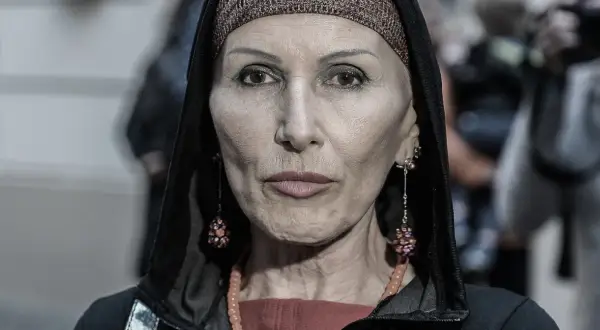 Kobieta w kapturze i ozdobnej opasce na głowie. Jej twarz, naznaczona kora wiek, opowiada historię. Długie kolczyki i naszyjnik dodają jej charakteru.