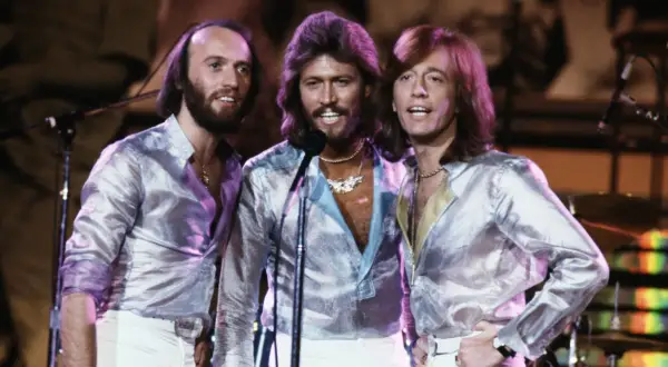 Bracia Bee Gees w błyszczących koszulach i białych spodniach, śpiewający na scenie.