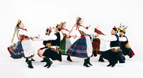 Tancerze w barwnych strojach ludowych, w tym w tradycyjnych strojach kujawiaków, tworzą taneczny krąg.