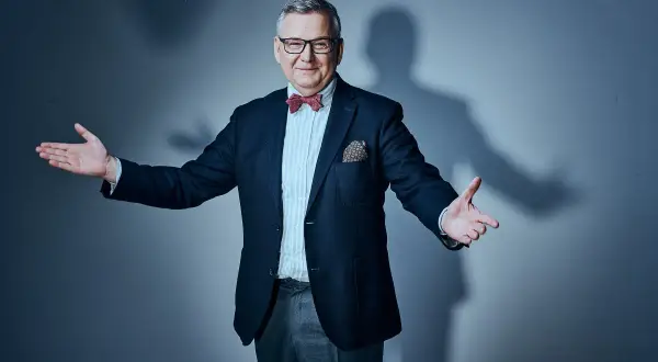 Artur Andruś, w eleganckim garniturze i muszce, gestykuluje, jakby opowiadał o swojej rodzinie.