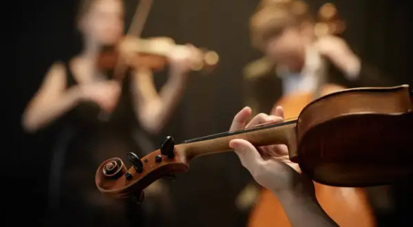 Dłoń trzymająca gryf skrzypiec, w tle dwie kobiety grające na instrumentach smyczkowych. To preludium do pięknej muzyki.