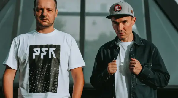 Dwóch członków Paktofoniki, jeden w białej koszulce z logo, drugi w czarnej kurtce i czapce z daszkiem.