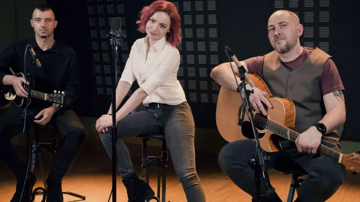 Zespół wykonuje brathanki utwory. Dwóch mężczyzn gra na gitarach, a kobieta z rudymi włosami siedzi pośrodku.