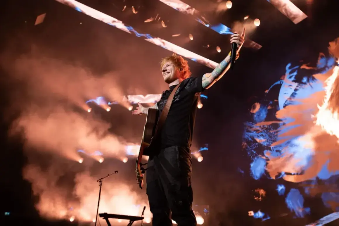 Ed Sheeran w Polsce, na scenie z gitarą, w otoczeniu dymu i świateł, pozdrawia publiczność.
