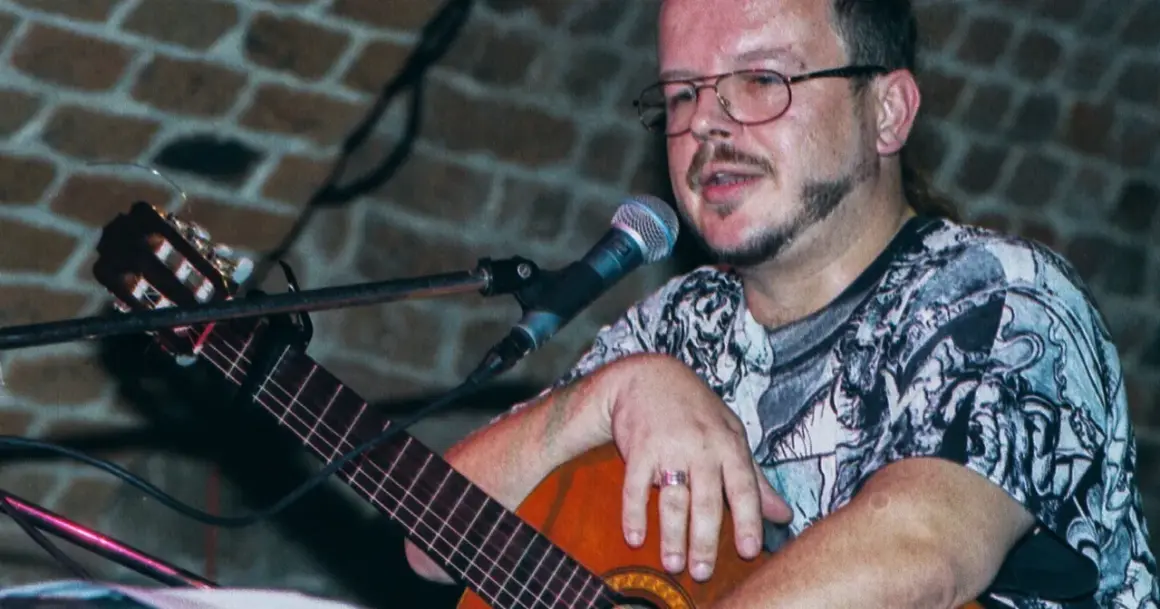 Jacek Kaczmarski z gitarą i mikrofonem, w okularach i wzorzystej koszulce. Prywatne chwile artysty.