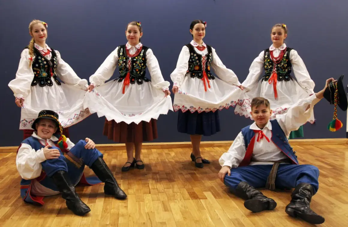 Dzieci w strojach ludowych, dziewczęta trzymają białe spódnice, chłopcy w niebieskich spodniach i kamizelkach, gotowi do tańca kołomyjka.