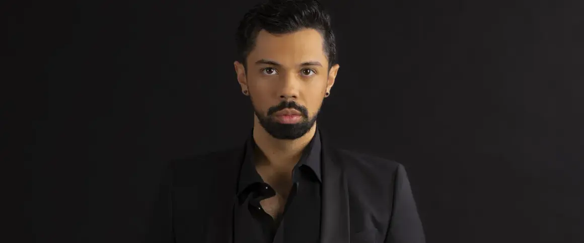 Mężczyzna z brodą i kolczykiem w uchu, ubrany w czarną koszulę i marynarkę. Wygląda jak młody opera singer.