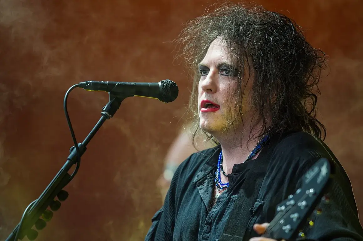 Robert Smith, the iconic wokalista The Cure, śpiewa do mikrofonu, z charakterystycznym makijażem i rozczochranymi włosami.