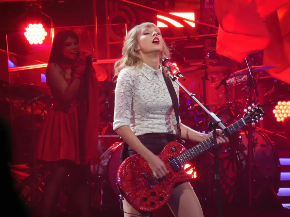 Taylor Swift gra na czerwonej gitarze podczas koncertu. W tle widać perkusję i inną wokalistkę. To może być fragment trasy promującej jej taylor swift albumy.