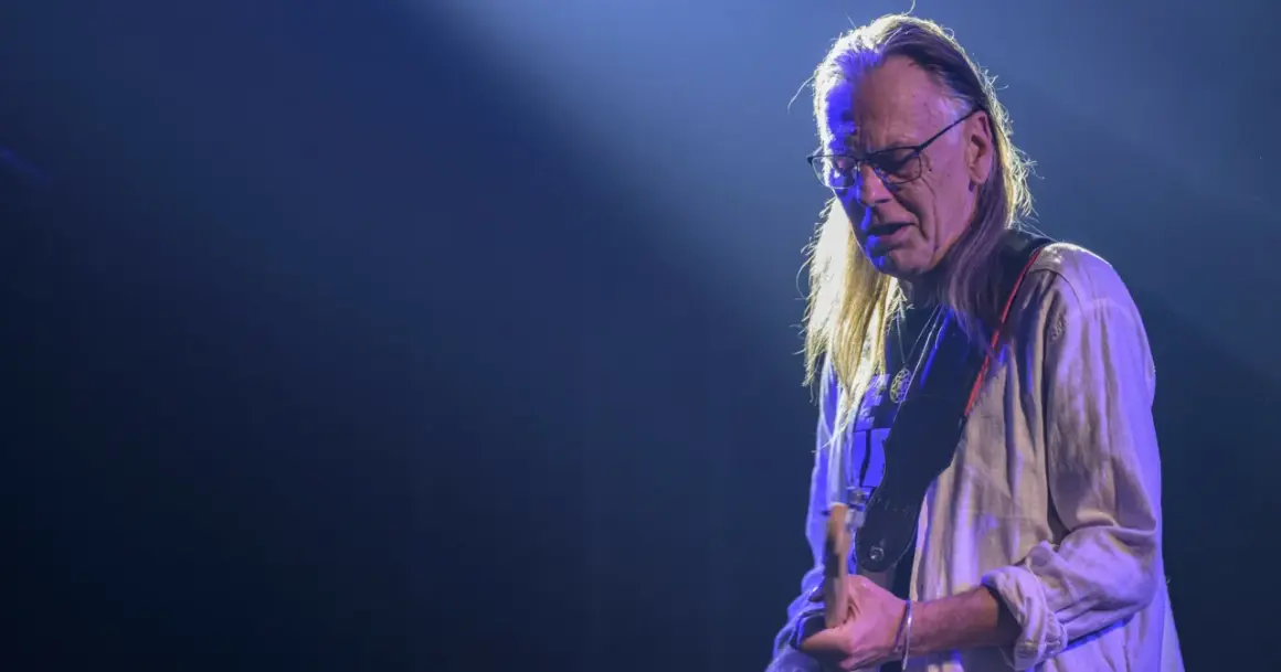 Muzyk z zespołu Lipińskiego w okularach i z długimi włosami, grający na gitarze podczas koncertu.