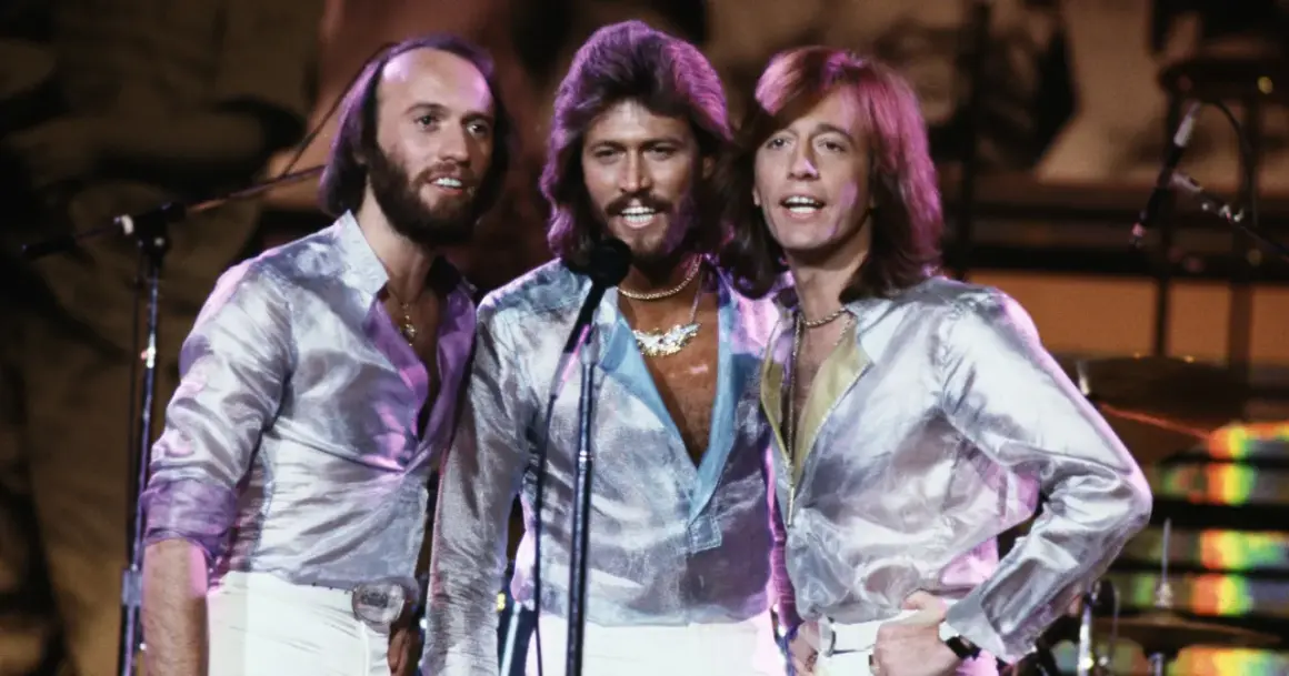 Bracia Bee Gees w błyszczących koszulach i białych spodniach, śpiewający na scenie.