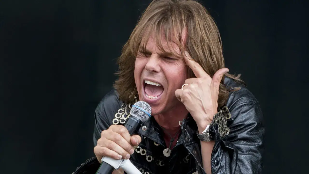 Joey Tempest, wokalista Europe, śpiewa z pasją, wskazując palcem na skroń.
