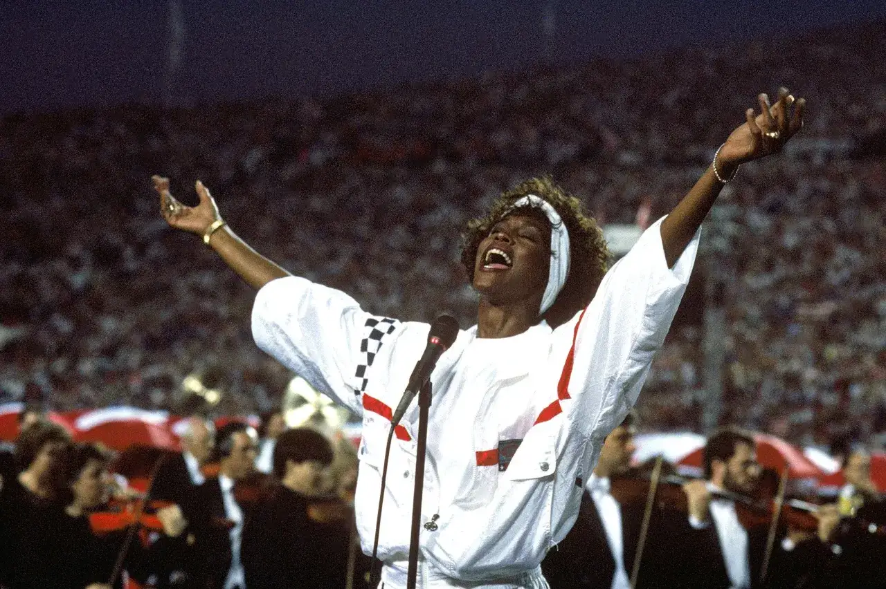 Whitney Houston w wieku 26 lat, z rękami w górze, śpiewa przed tłumem.
