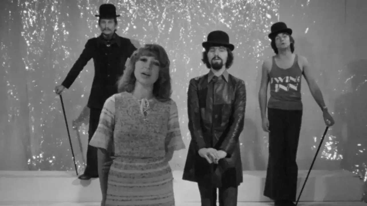 Ewa Wanat i jej zespół Novi Singers w teatralnych strojach, z melonikami i laseczkami, na tle błyszczącej scenografii.