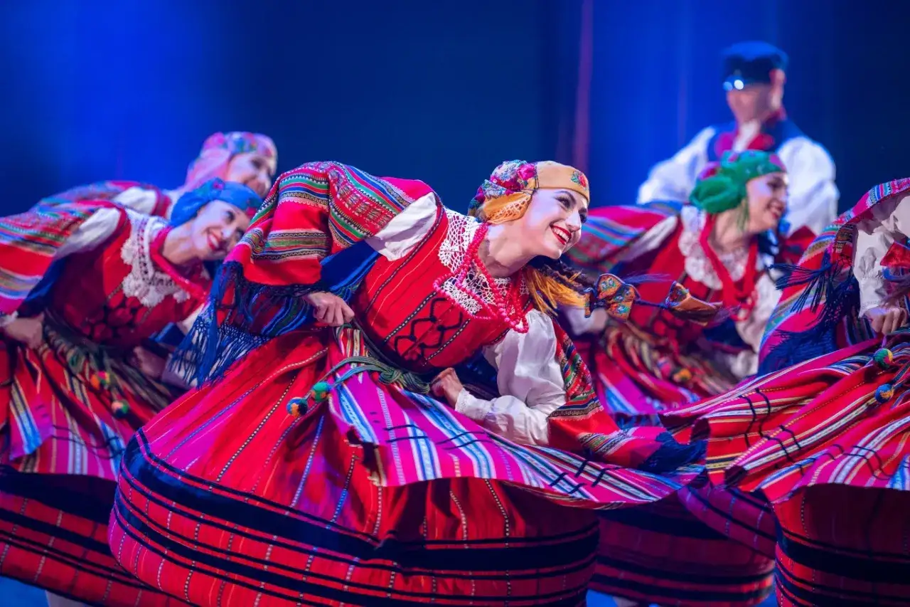 Tancerki w barwnych strojach ludowych, wirujące w radosnym tańcu. Żywy folklor na scenie.