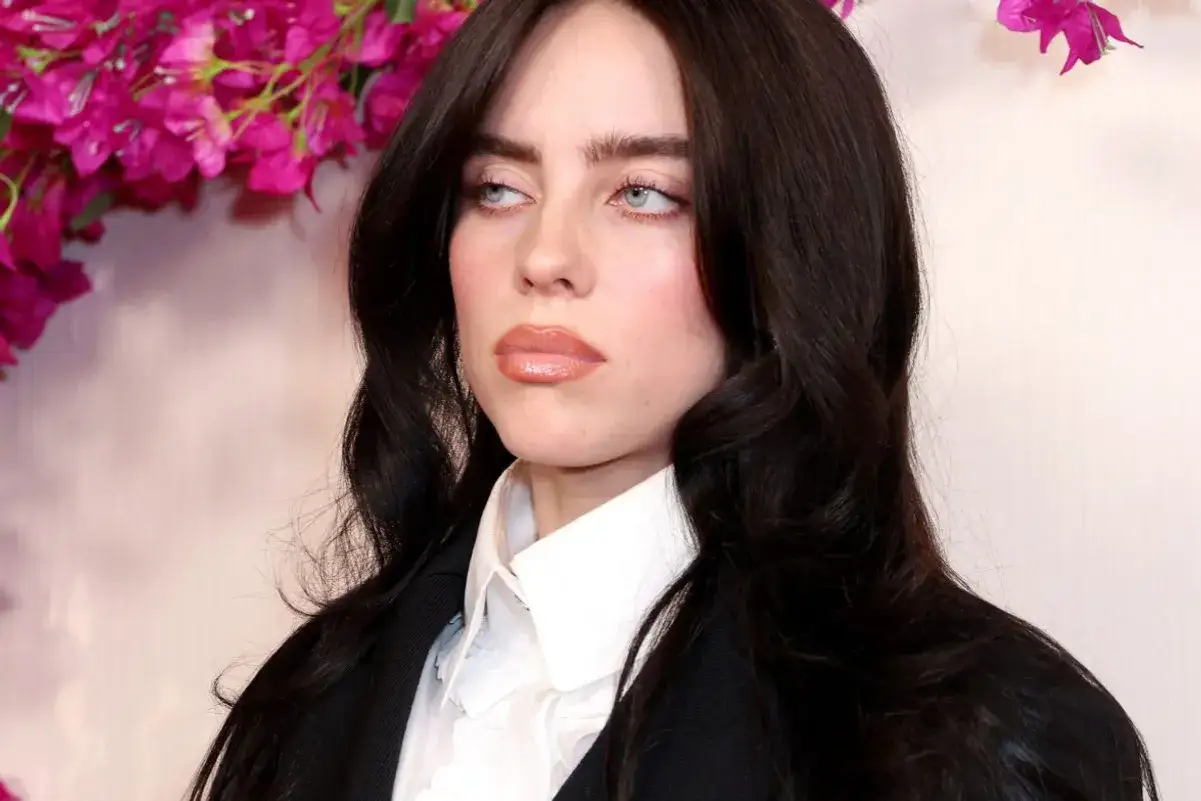 Billie Eilish w czarnej marynarce i białej koszuli, z burzą ciemnych włosów. Jej spojrzenie sugeruje głębię emocji, jak w jej utworach.