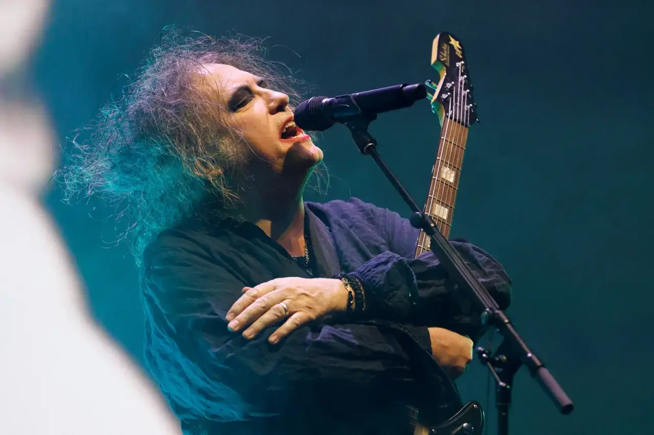 Robert Smith, the wokalista The Cure, śpiewa do mikrofonu z gitarą na scenie, jego włosy rozwiewane są przez światła.