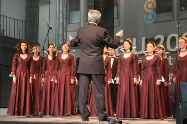 Chór kobiecy w bordowych sukniach, dyrygowany przez mężczyznę w garniturze. W tle logo "European Capital of Culture".