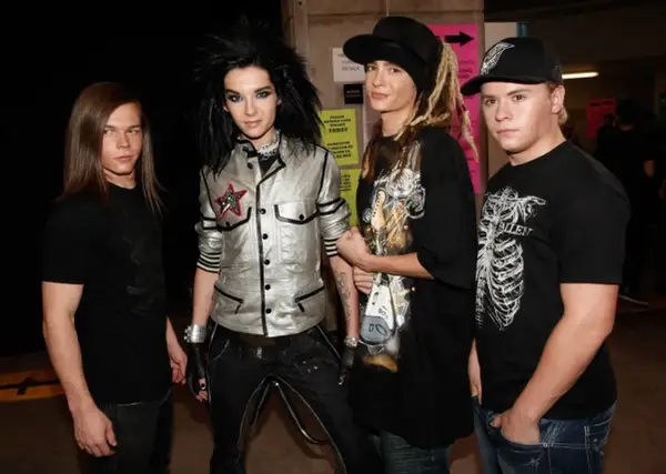 Członkowie Tokio Hotel pozują do zdjęcia. Bill Kaulitz w błyszczącej kurtce, Tom Kaulitz w czapce i z dredami, a obok nich Georg Listing i Gustav Schäfer.