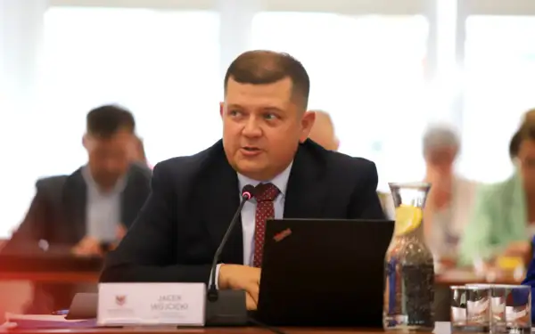 Jacek Wójcicki, siedząc przy stole, przemawia. Jego wzrost jest przeciętny, a na stole leży laptop i woda z cytryną.