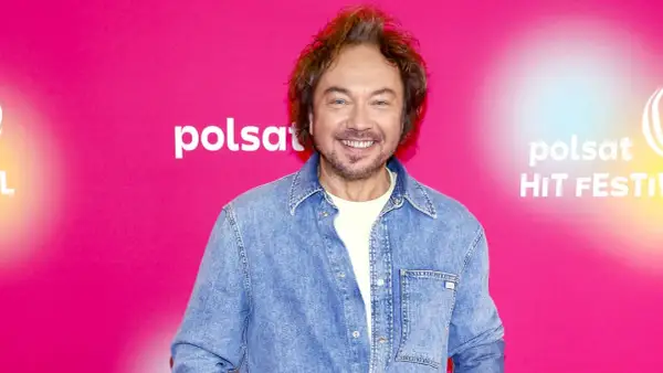 Mietek Szczęśniak, uśmiechnięty, w dżinsowej koszuli, na tle różowego banera z logo Polsat Hit Festiwal.