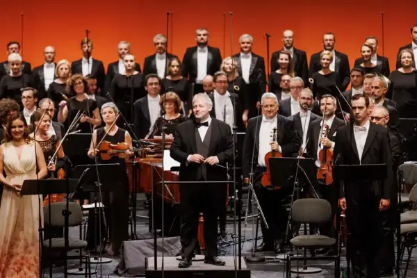 Orkiestra i chór przygotowują się do wykonania **Moniuszko opery**. Dyrygent stoi na podwyższeniu, gotowy do rozpoczęcia.