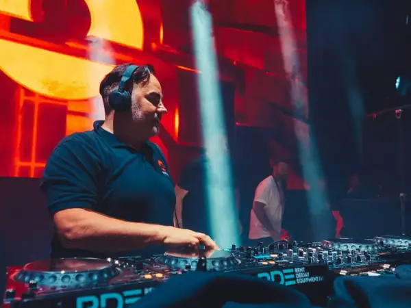 DJ Kris w akcji, miksuje muzykę na żywo. W tle pulsujące światła i tłum.