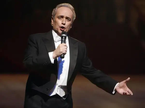José Carreras, hiszpański tenor, w eleganckim smokingu śpiewa do mikrofonu, gestykulując.