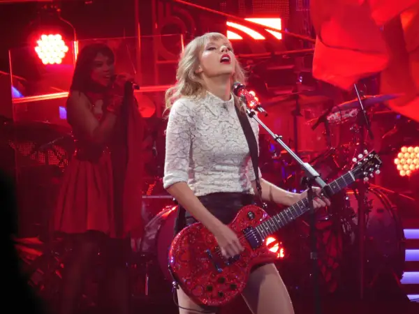 Taylor Swift gra na czerwonej gitarze podczas koncertu. W tle widać perkusję i inną wokalistkę. To może być fragment trasy promującej jej taylor swift albumy.