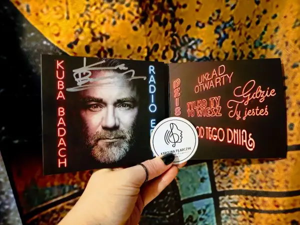 Kuba Badach z autografem na okładce płyty. Neonowe napisy "Dziś układ otwarty", "Tylko ty to wiesz", "Gdzie ty jesteś", "Od tego dnia".
