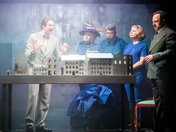 Aktorzy na scenie, prezentujący polskie musicale, gestykulują przy makiecie miasta.