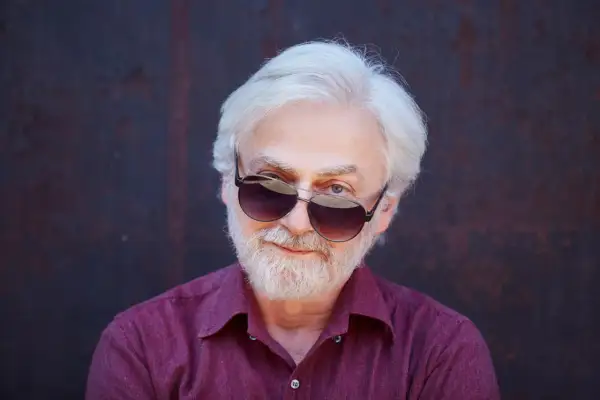 Krystian Zimerman w okularach przeciwsłonecznych, z siwą brodą i włosami, w bordowej koszuli.
