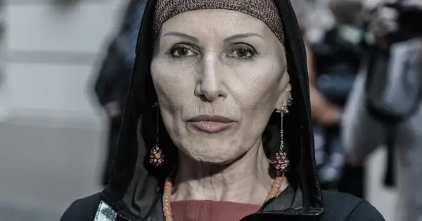 Kobieta w kapturze i ozdobnej opasce na głowie. Jej twarz, naznaczona kora wiek, opowiada historię. Długie kolczyki i naszyjnik dodają jej charakteru.